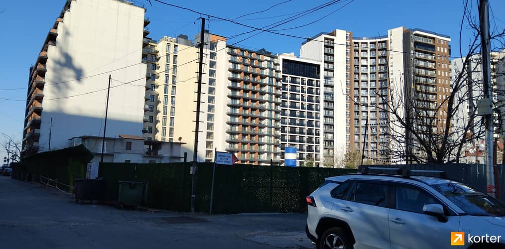 Реальная фотография фасада многоэтажного жилого комплекса с улицы, на переднем плане виден автомобиль и ограждение.